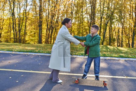 mère et fils dans le parc, joyeuse femme afro-américaine tenant la main avec un garçon sur un penny board, automne