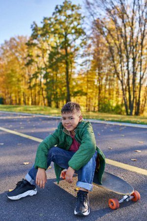 Foto de Lindo afroamericano chico sentado en penny board, parque de otoño, temporada de otoño, ropa de abrigo, ropa de abrigo - Imagen libre de derechos