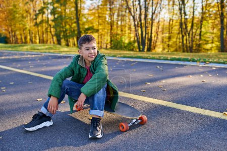 chico afroamericano pensativo en ropa de abrigo sentado en penny board, parque de otoño, temporada de otoño