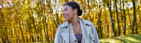 cheerful african american woman in trench coat walking in autumn park, fashion outfit, banner