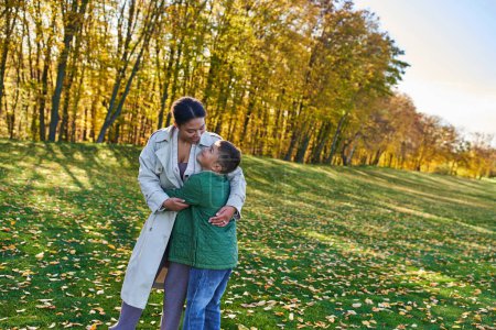 mujer afroamericana feliz abrazándose con hijo, de pie sobre hierba con hojas doradas, otoño