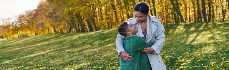 happy african american woman hugging with son, standing on grass with golden leaves, autumn, banner