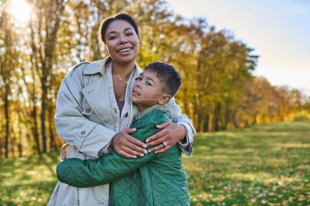 Mutterliebe, Freude, afrikanisch-amerikanische Frau umarmt niedlichen Sohn, steht neben goldenen Blättern, Herbst