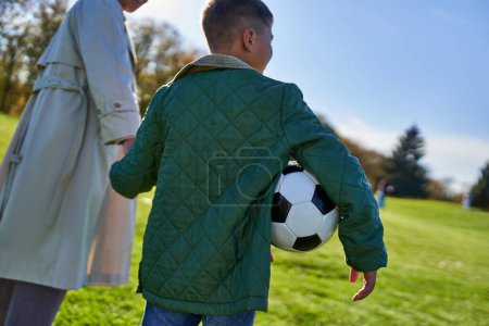 chico afroamericano sosteniendo el fútbol, caminando con mamá en el campo, tomados de la mano, caída, primer plano