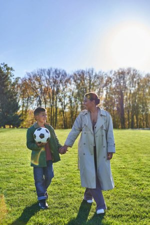 afrikanisch-amerikanischer Junge hält Fußball, geht mit Mama auf dem grünen Feld, hält Händchen, Herbst