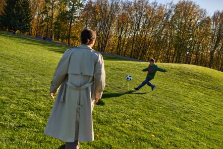 lindo chico afroamericano jugando fútbol cerca de la madre en el campo verde, fútbol, otoño, temporada de otoño