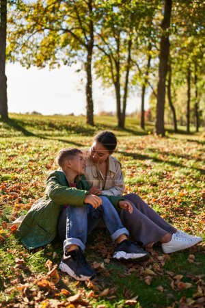 amor maternal, mujer afroamericana feliz e hijo sentado en la hierba con hojas doradas, otoño