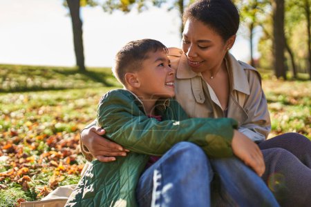 amour maternel, gaie femme afro-américaine et son fils étreignant et assis près des feuilles d'or, automne