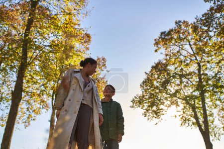 african american mother and child in outerwear holding hands near trees in autumn park, fall season