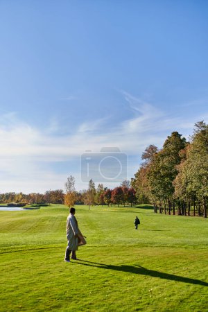 african american woman in trench coat looking at son running on green grass, trees, nature