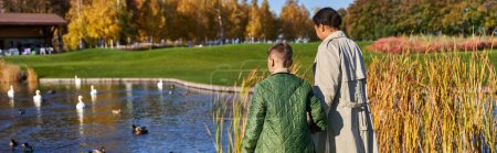 back view of mother and son in outerwear walking together near lake with swans and ducks, banner