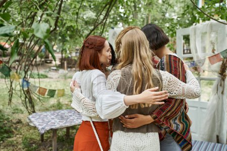 smiling multiethnic women in boho clothes hugging while standing outdoors in retreat center