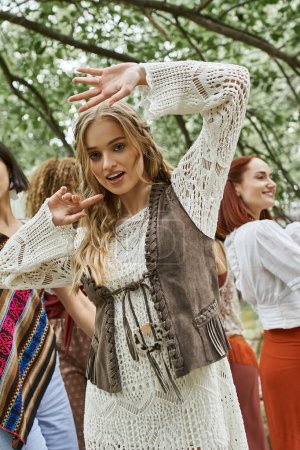 Young blonde woman in boho outfit looking at camera and dancing near friends in retreat center