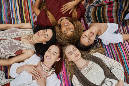 top view of smiling multiethnic women in boho outfits lying on blanket in retreat center