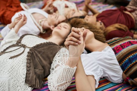 blurred multiethnic women holding hands while lying on blanket outdoors in retreat center