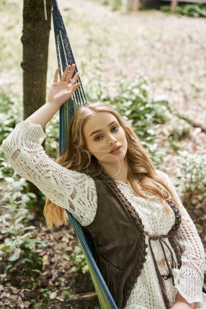 Photo for Young blonde woman in boho outfit looking at camera on hammock in retreat center - Royalty Free Image