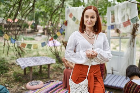 positive redhead woman in boho outfits looking at camera while standing outdoors in retreat center