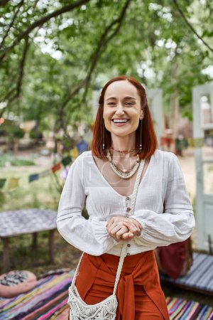 portrait of excited redhead woman in boho outfit looking at camera outdoors in retreat center