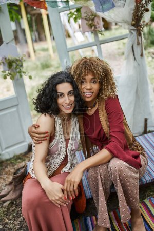 smiling interracial girlfriends in boho outfits hugging and looking at camera in retreat center