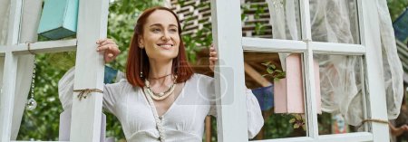 joyful and stylish redhead woman standing near doors outdoors in retreat center, banner