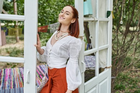 positive and trendy redhead woman in boho styled outfit standing outdoors in retreat center