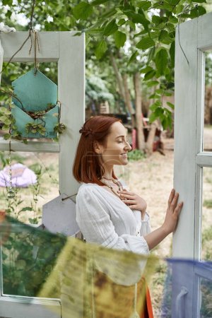 pleased and trendy redhead woman in boho clothes standing outdoors in retreat center