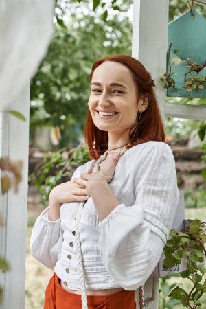 pleased redhead woman in stylish outfit looking at camera in patio in retreat center