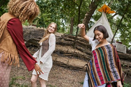 young and interracial girlfriends in boho outfits dancing on lawn in retreat center