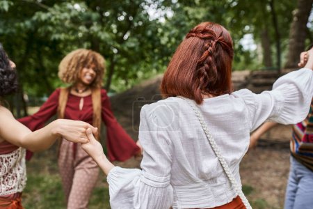 redhead woman in boho clothes holding hands of blurred multiethnic friends in retreat center