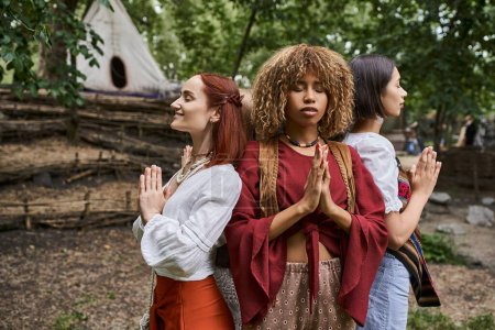 multiethnic women with closed eyes doing praying hands gesture outdoors in retreat center
