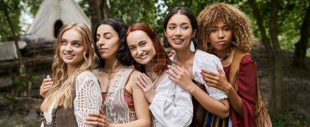 positive multiethnic women in boho outfits hugging outdoors in retreat center, banner