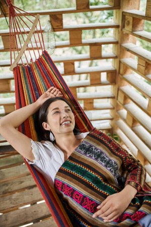 carefree and stylish woman smiling while relaxing in hammock in retreat center