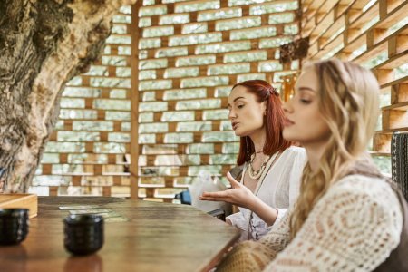 woman pointing at tarot cards near blurred girlfriend in cottage on retreat center