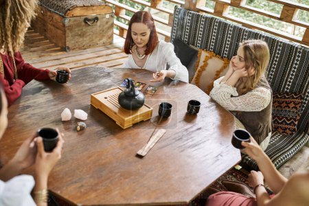 Frau liest Tarot-Karten in der Nähe multiethnischer Freundinnen und Tee auf Holztisch im gemütlichen Haus