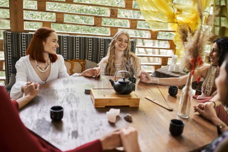 joyful girlfriends holding hands and meditating near teapot and cups on wooden table in cottage