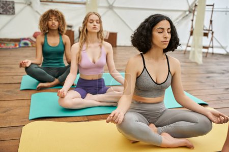 multiethnic women in sportswear sitting on yoga mats and meditating in lotus pose, healthy lifestyle