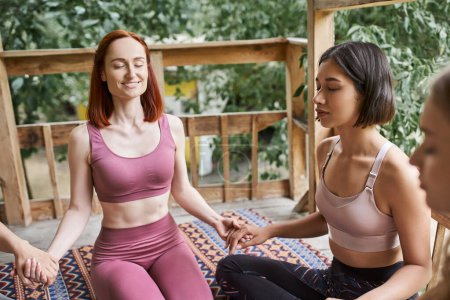 cheerful woman holding hands with girlfriends and meditating with closed eyes in retreat center