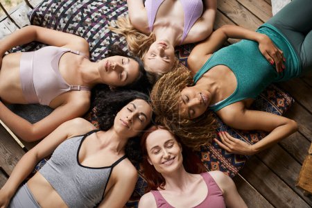 Draufsicht auf unbeschwerte multiethnische Freundinnen, die mit geschlossenen Augen auf Kissen meditieren, Ruhe
