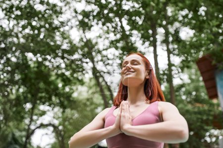 fröhliche rothaarige Frau meditiert mit geschlossenen Augen und betenden Händen im Park, Rückzugskonzept für Frauen