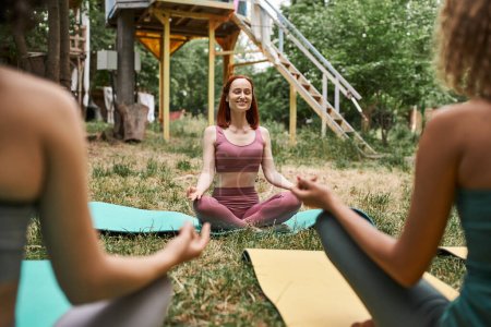 freudige Frau mit geschlossenen Augen meditiert im Park des Exerzitienzentrums neben verschwommenen Freundinnen