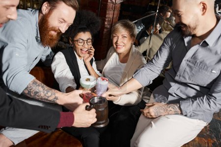 happy women and tattooed men clinking glasses of cocktails in bar, multiethnic friends having fun