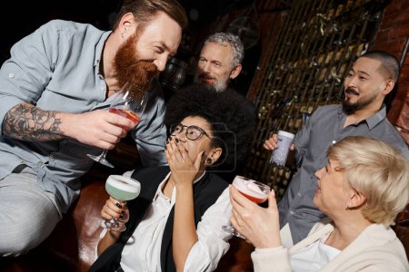lachende afrikanisch-amerikanische Frau deckt Mund mit der Hand in der Nähe multiethnischer Freunde mit Getränken in der Bar ab