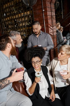 bearded tattooed men talking near women drinking cocktails in bar, leisure of multiethnic colleagues