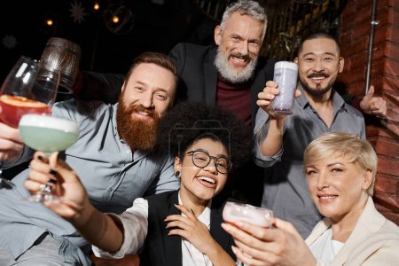 collègues multiethniques joyeux avec cocktail regardant la caméra dans le bar, temps heureux après le travail