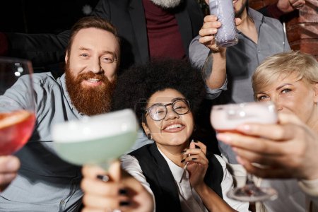 hombre barbudo feliz y mujer afroamericana con cócteles mirando a la cámara cerca de colegas en el bar