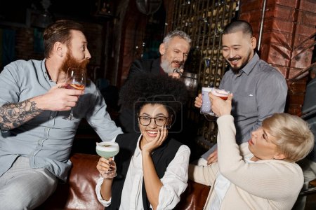 joyful african woman looking at camera near multiethnic colleagues clinking glasses in cocktail bar