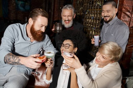 Bärtiger tätowierter Mann mit Brille und multiethnischen Frauen neben lächelnden Arbeitskollegen in Cocktailbar