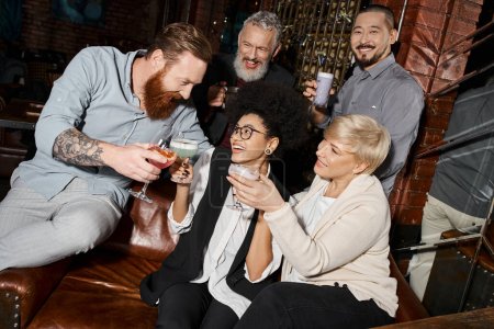 Bärtiger tätowierter Mann mit Brille und multiethnischen Frauen neben lächelnden Arbeitskollegen in Cocktailbar
