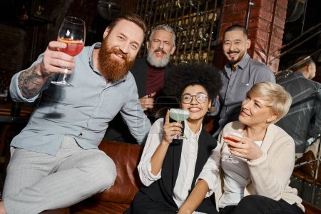 bärtiger tätowierter Mann mit Cocktail blickt in die Kamera in der Nähe multiethnischer Freunde, die sich in einer Bar amüsieren