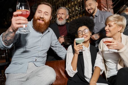 heureux tatoué et barbu homme regardant cocktail près de collègues multiethniques avec des lunettes dans le bar